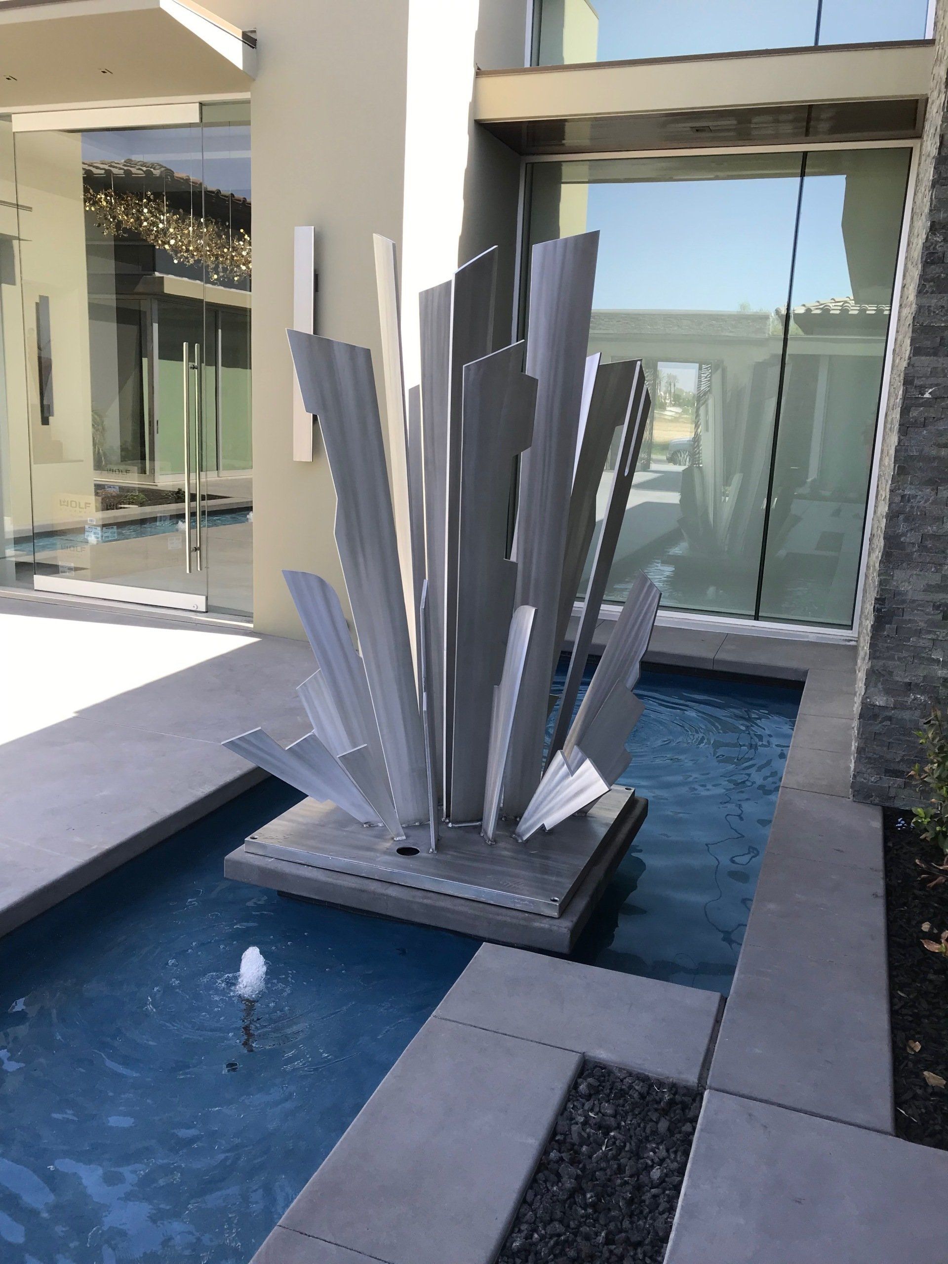 Modern outdoor water fountain with abstract metal sculpture; rectangular pool, glass windows.