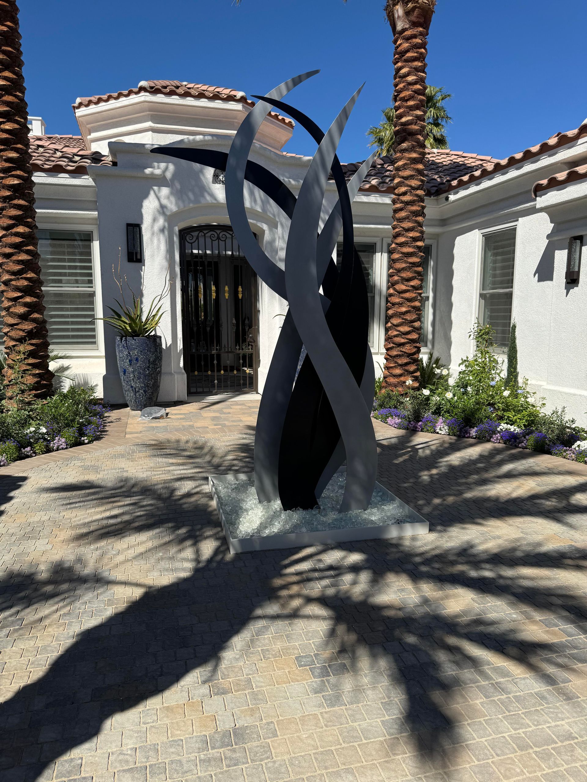 Metal sculpture in front of a white house, palm trees in the background, bright sunlight casting shadows.