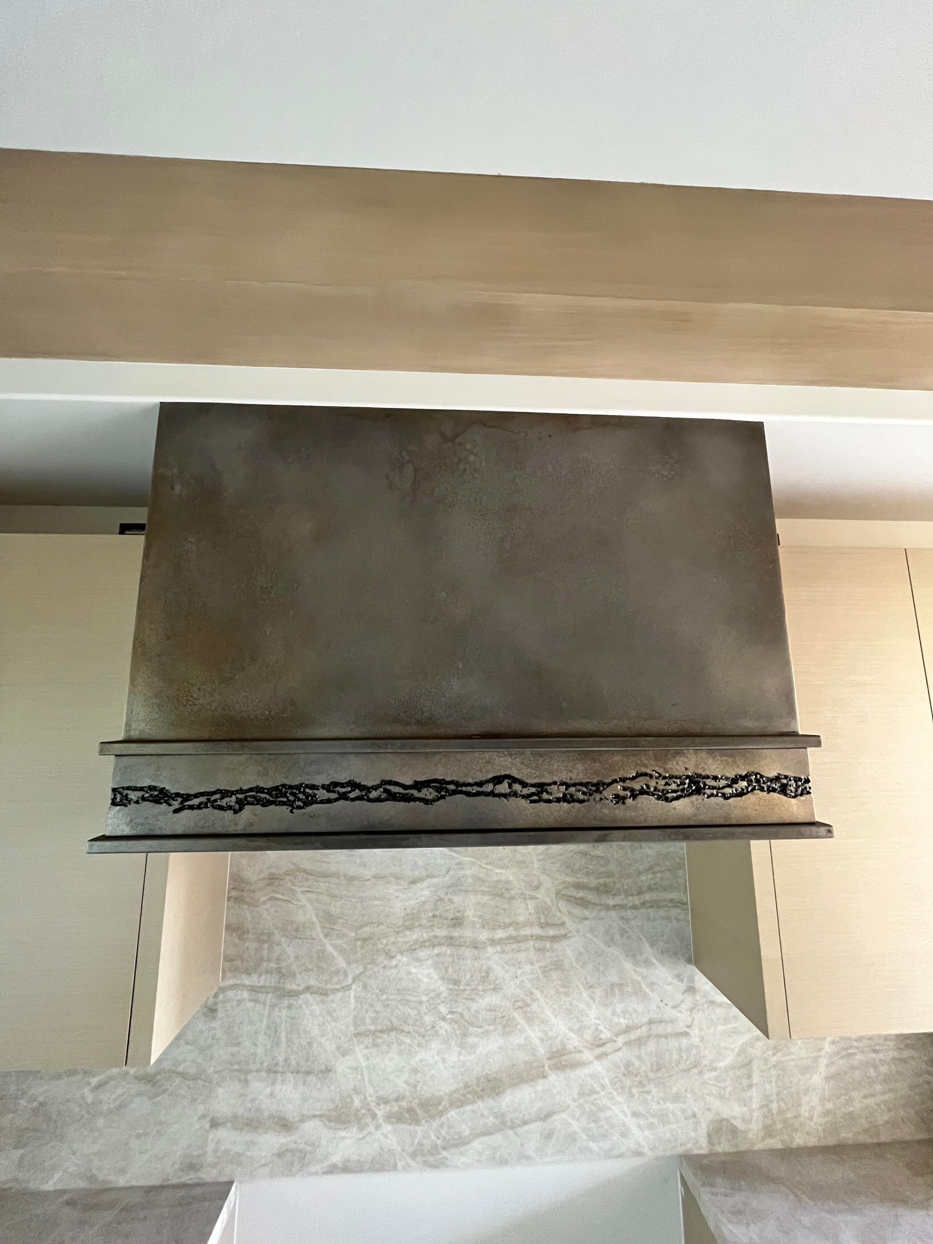 Stainless steel range hood with decorative welded detail, mounted above a tiled backsplash.