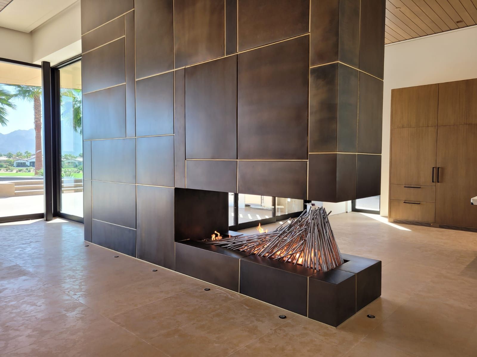 Modern fireplace with dark metal panels and sculptural fire feature; open to a bright outdoor view.