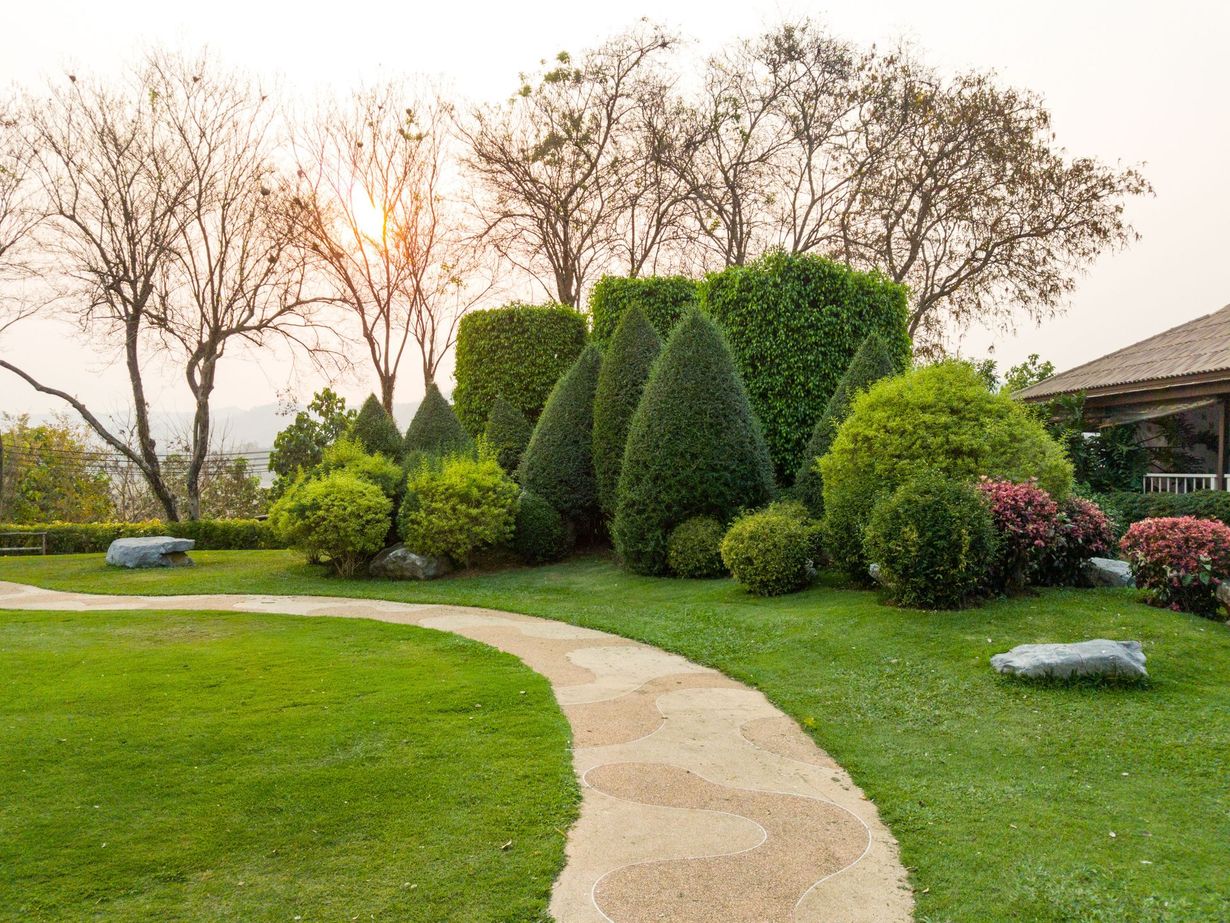 Stone path winds through a green, manicured garden with sculpted bushes and trees; sunlight.