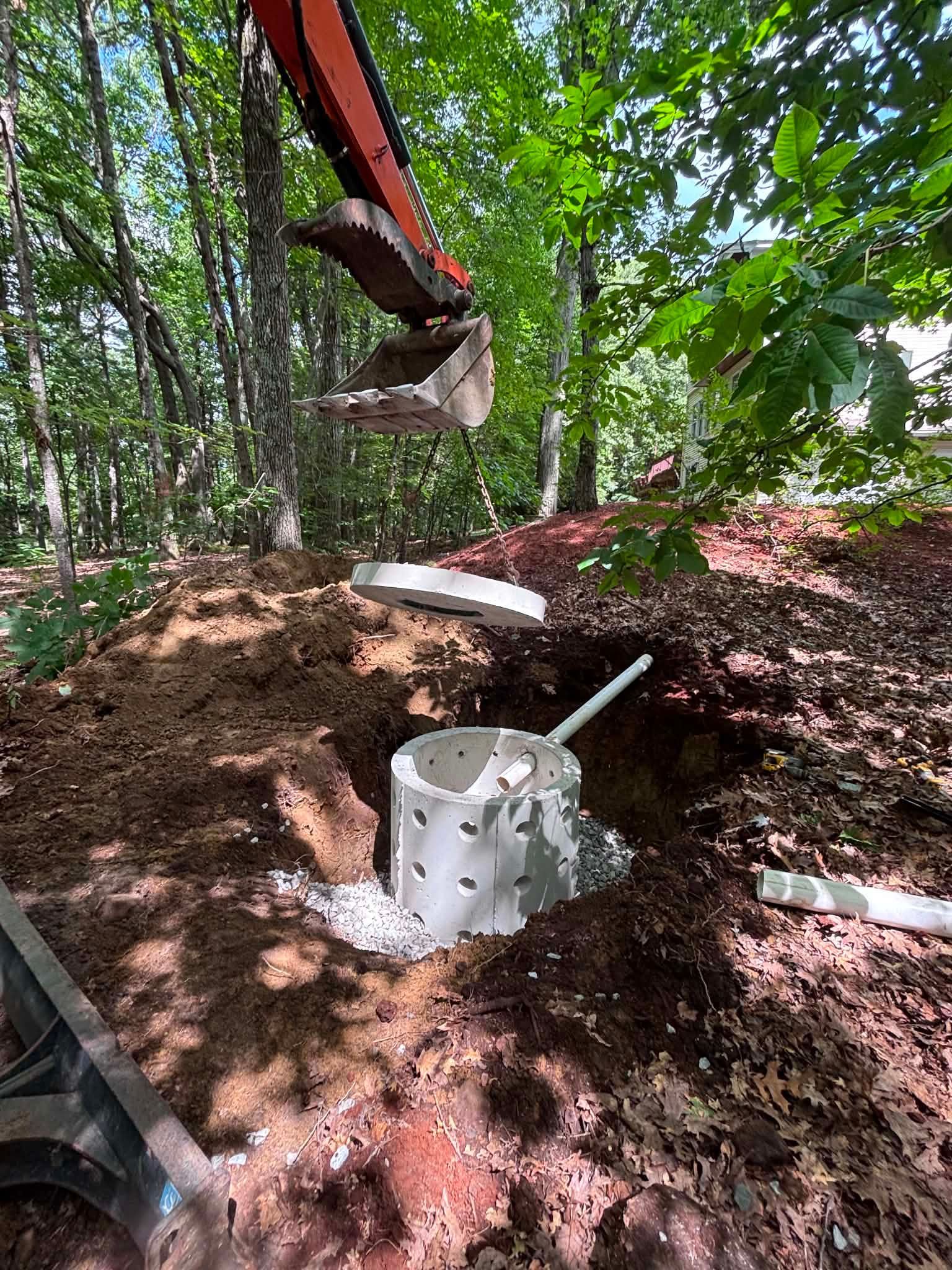 Excavator arm installing a white pipe fitting in a wooded dirt trench