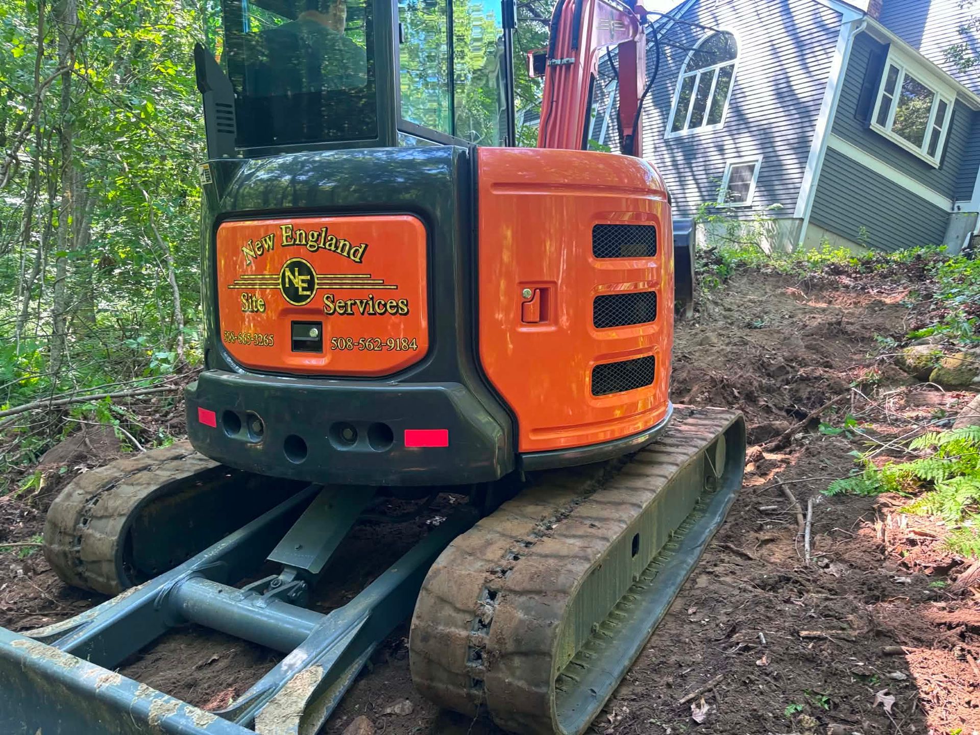 Orange tracked excavator on muddy ground in a wooded area, lifting logs near a small building