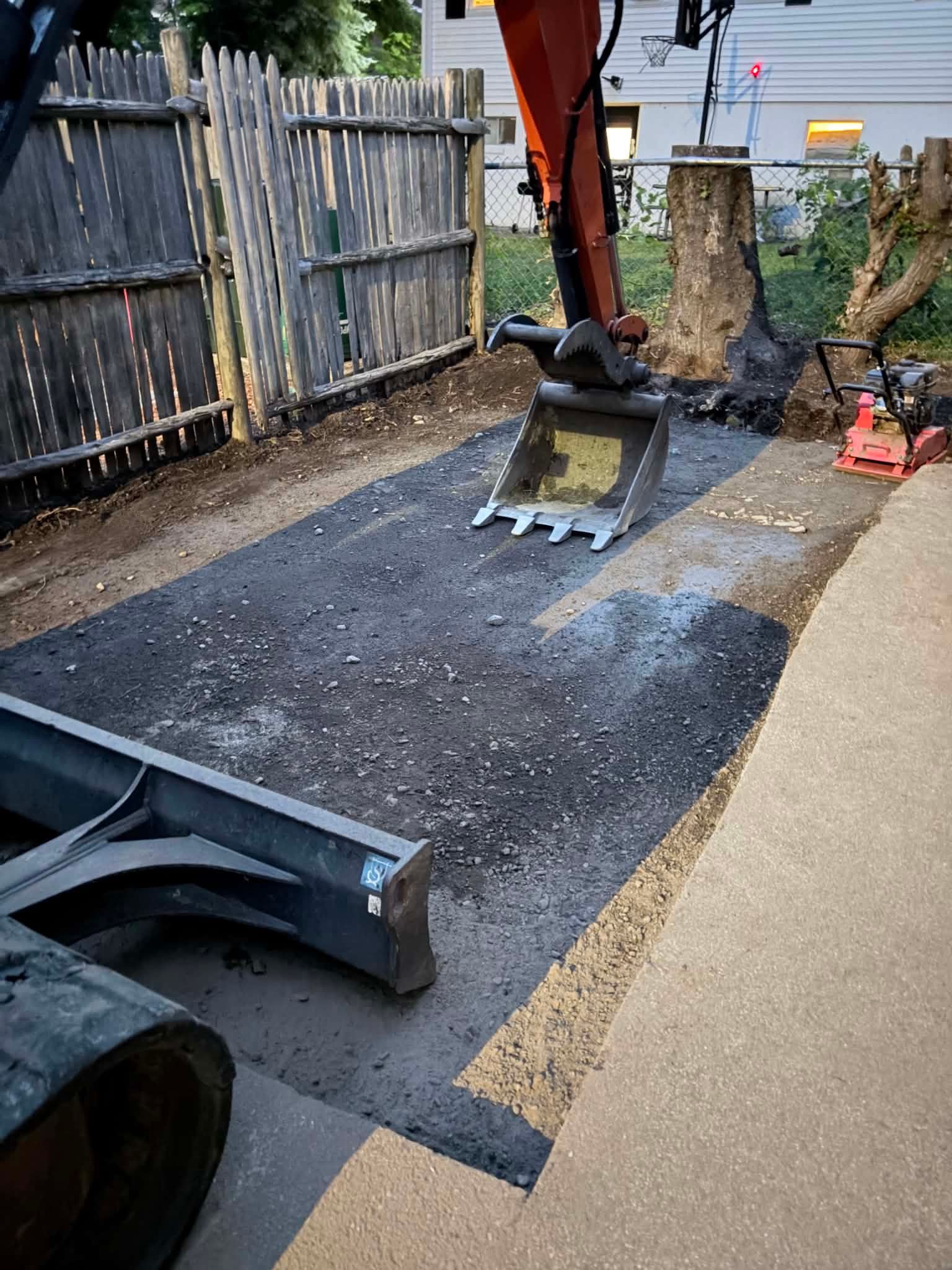 Construction machinery paving a driveway beside a wooden fence and tree stump.
