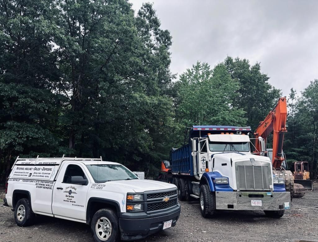 White utility truck and blue dump truck parked on gravel near trees and an orange excavator.