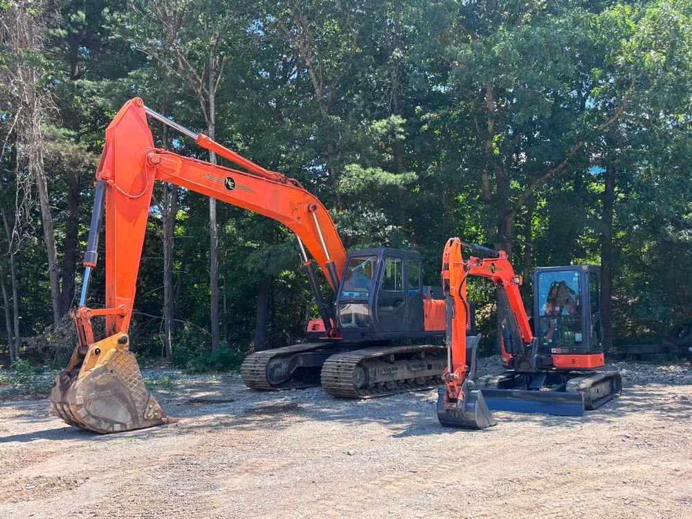 Two orange excavators parked on gravel in front of a wooded area.