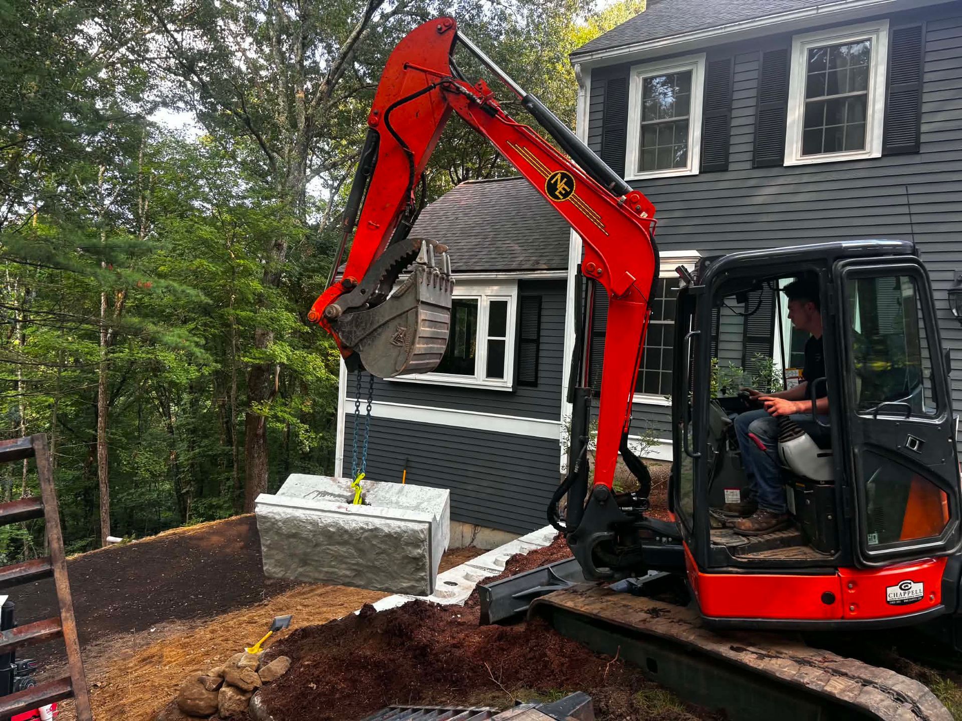 Red excavator beside a black house, lifting debris in a wooded yard.