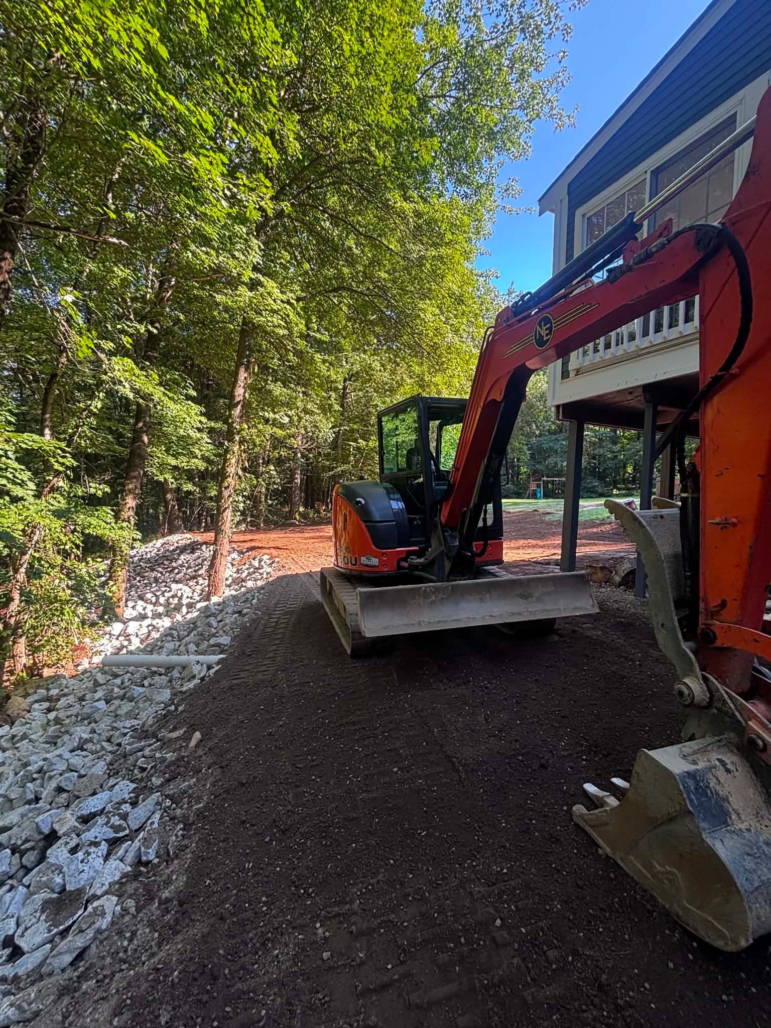 Orange excavator on gravel beside a wooded path under bright sunlight