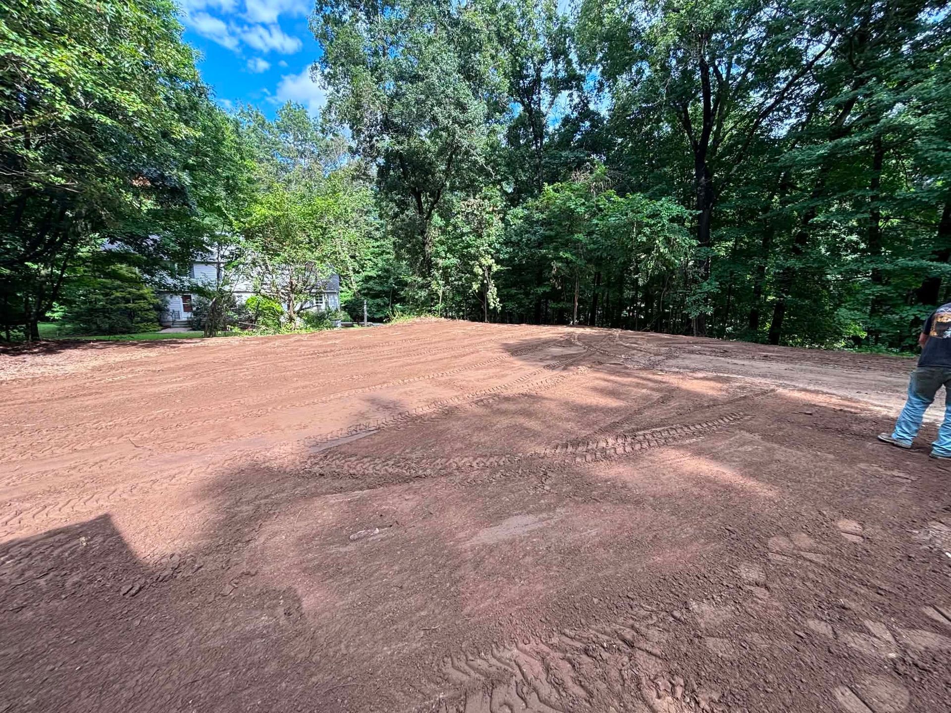 Freshly leveled dirt lot in a wooded park, with trees and blue sky overhead and a person standing at the edge
