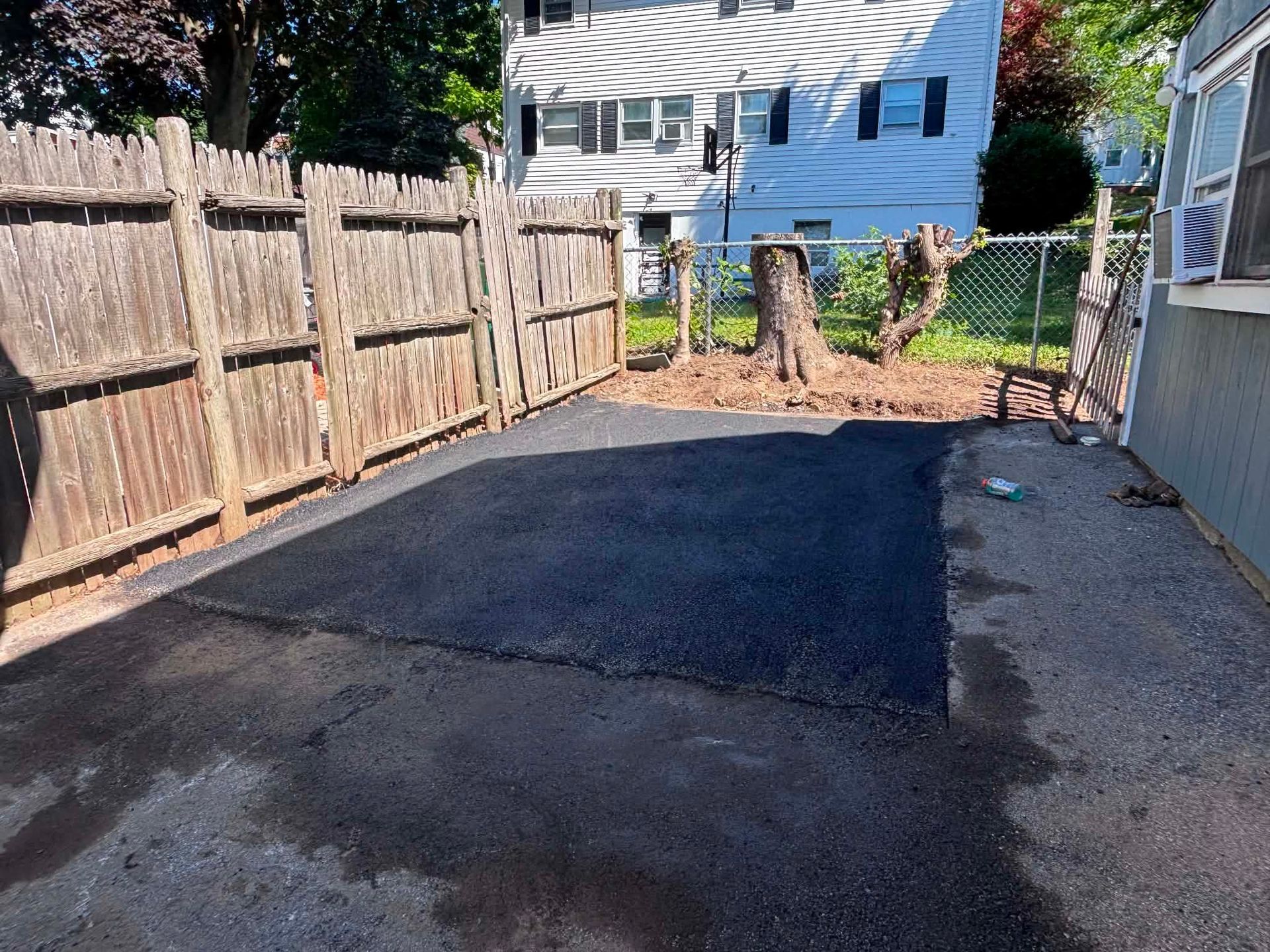 Freshly paved black asphalt driveway beside a wooden fence and house, with trees and a blue house in the background