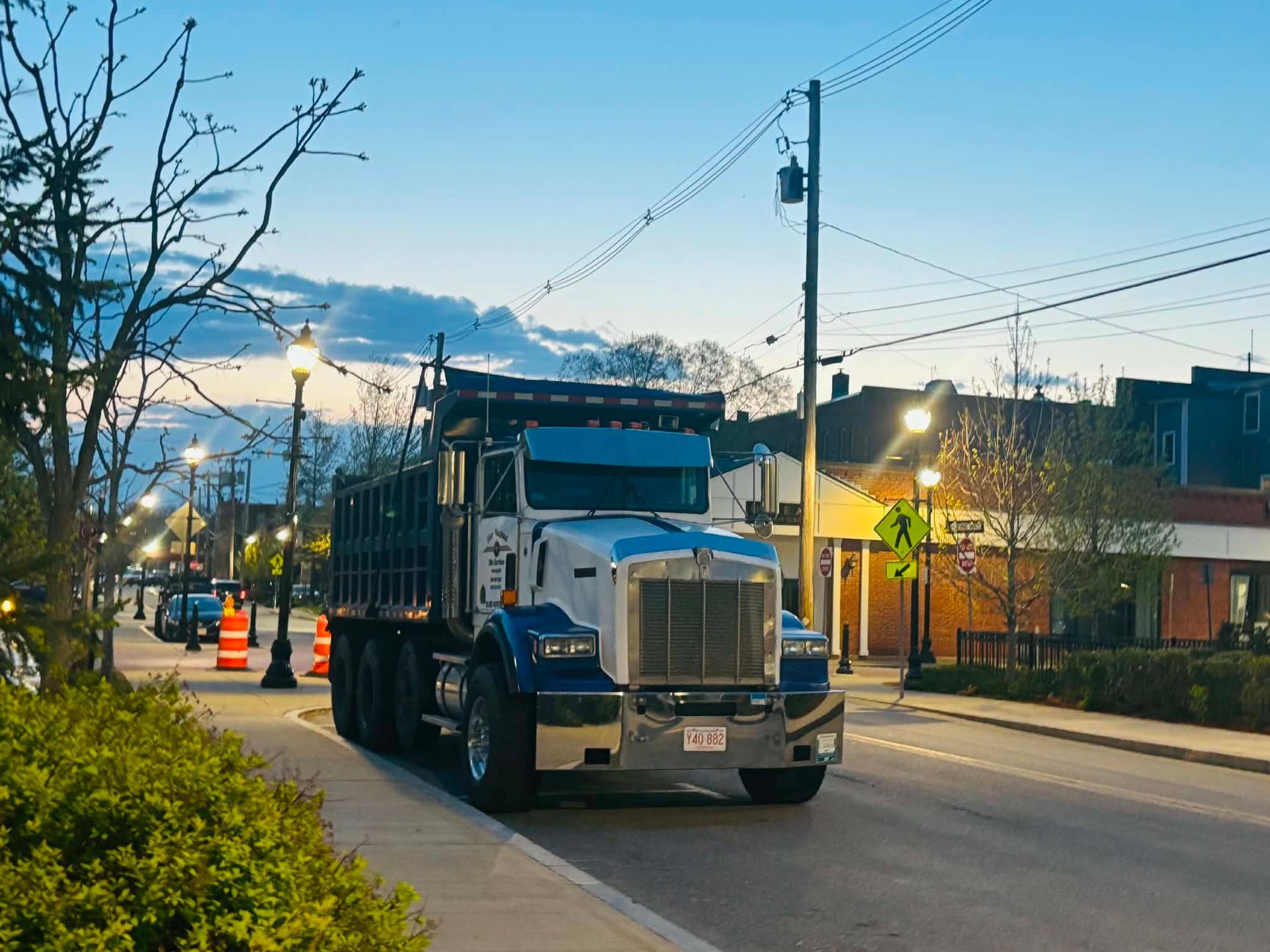Blue dump truck driving on a city street at dusk, with traffic cones and streetlights.