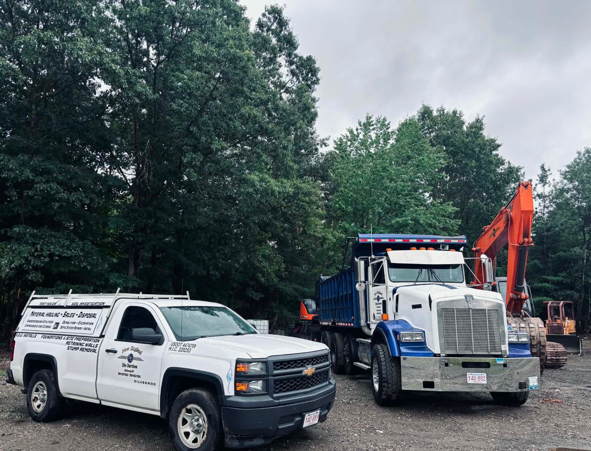 White service truck and blue dump truck parked on gravel by trees, with an excavator in the background