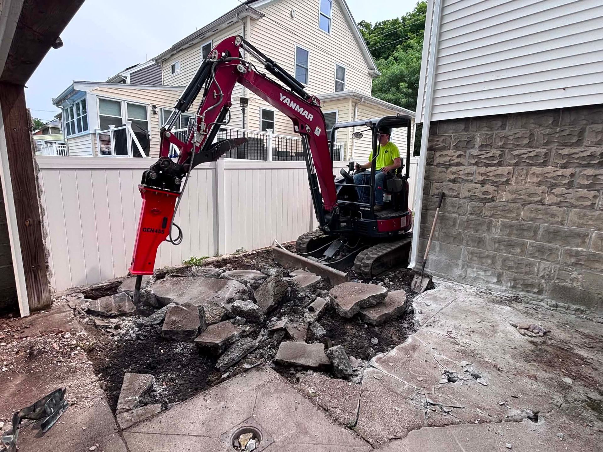 Mini excavator breaking up concrete beside a house and fence in a narrow yard