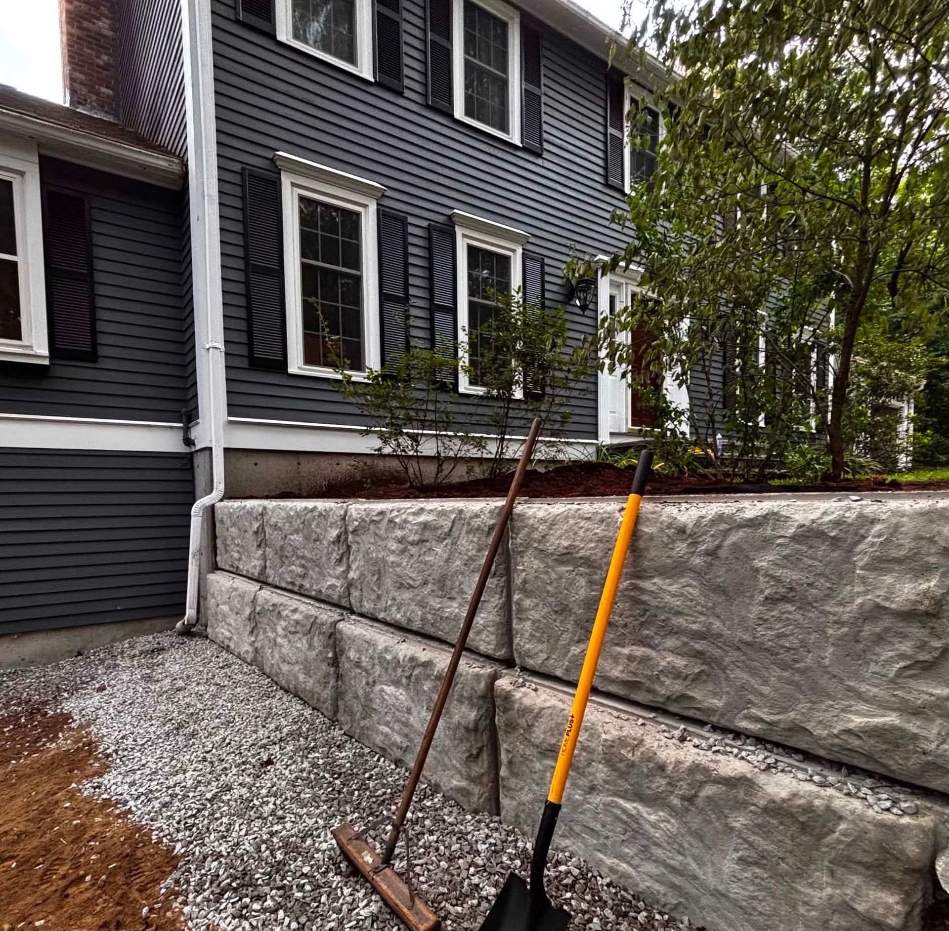 Two long-handled garden tools lean against a stone retaining wall beside a dark house.