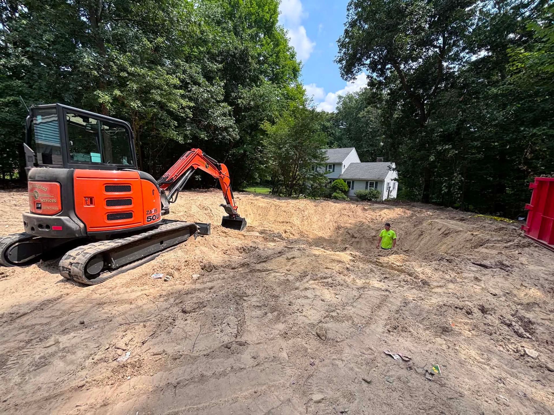 Orange excavator on a dirt lot near trees, with a worker in a neon vest and houses in the background