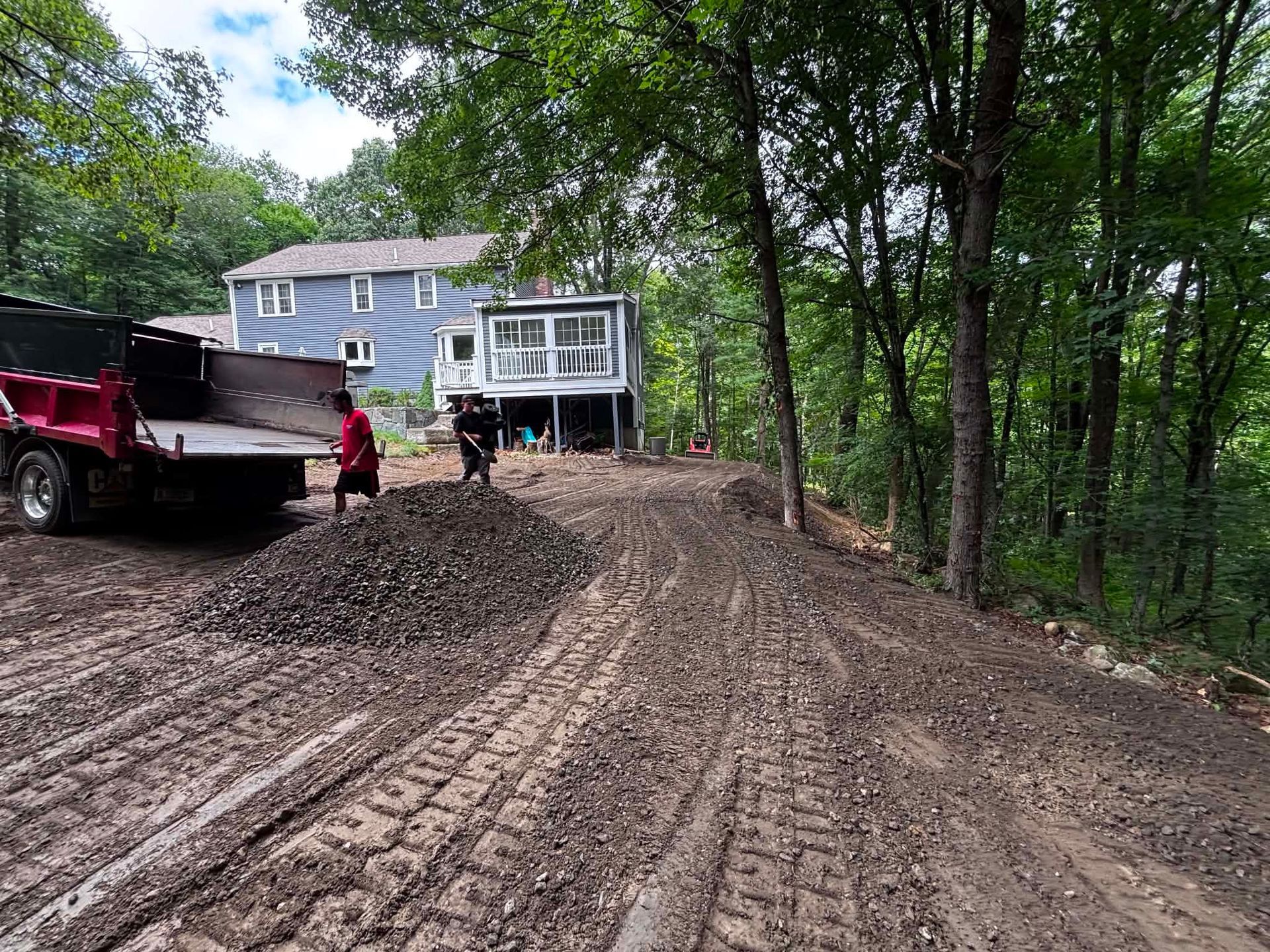 Mud-covered driveway with a dump truck, worker, and house in the background near trees