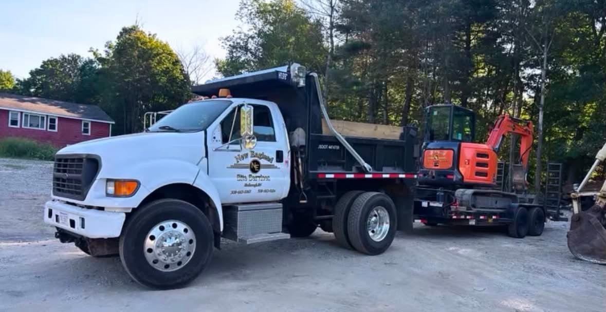 White dump truck hauling an orange excavator on a flatbed trailer in a wooded yard