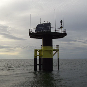 Offshore navigation tower with solar panels, black and yellow, against a cloudy sky.