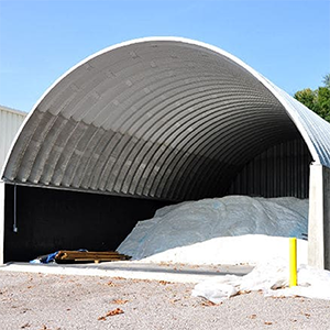 Curved metal building, housing a large pile of white salt; outdoors on a gravel surface.