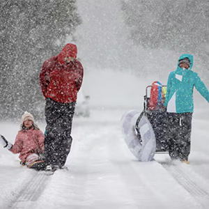 Family sledding in heavy snowfall on a snowy road; one child on the ground, others standing by a sled cart.