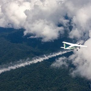 Airplane spraying a white plume over a green forest under a cloudy sky.