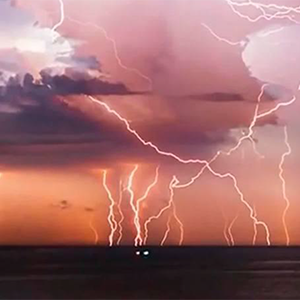 Lightning strikes over an ocean, illuminating the sky in shades of pink and orange during a storm.