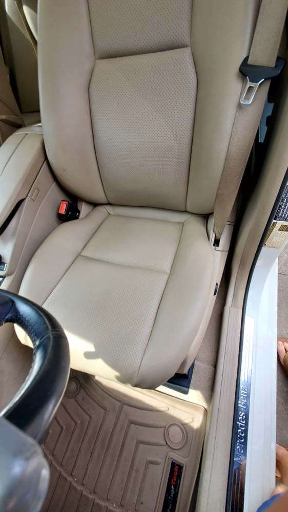 Beige leather car seat with seatbelt buckled, part of the steering wheel, and a floor mat.