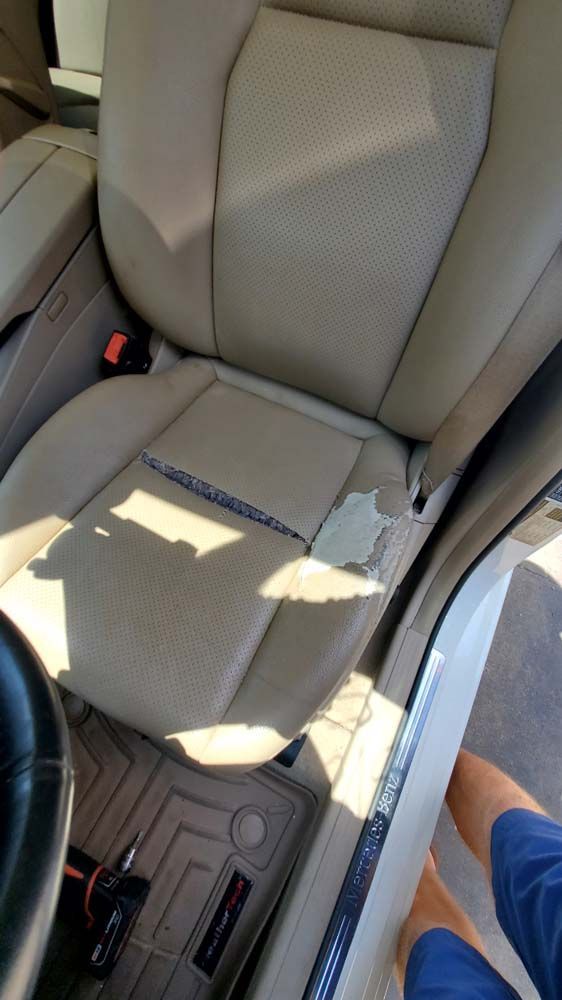 Beige car seat with ripped leather; part of a leg and steering wheel are visible.