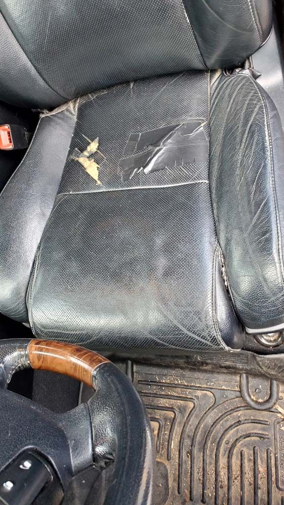 Damaged black leather car seat with torn sections, next to a steering wheel and floor mat.