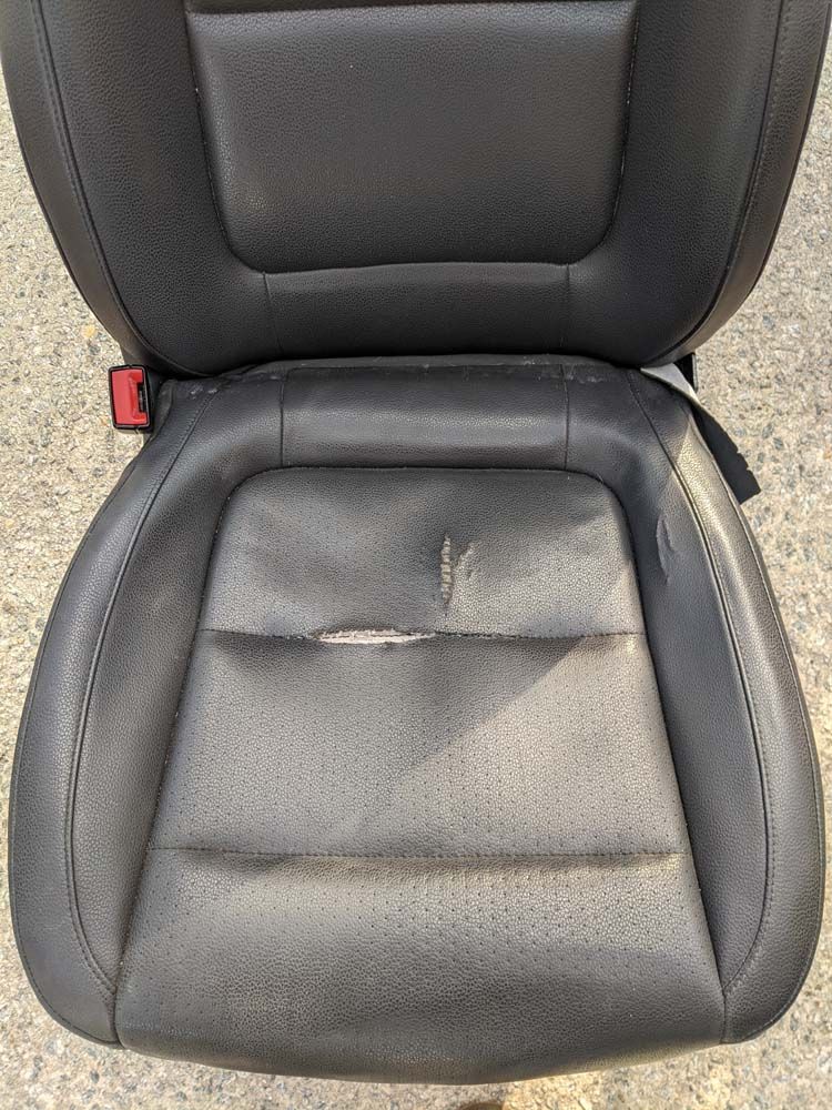 Black car seat with worn leather, visible seam and puncture on the seat cushion.