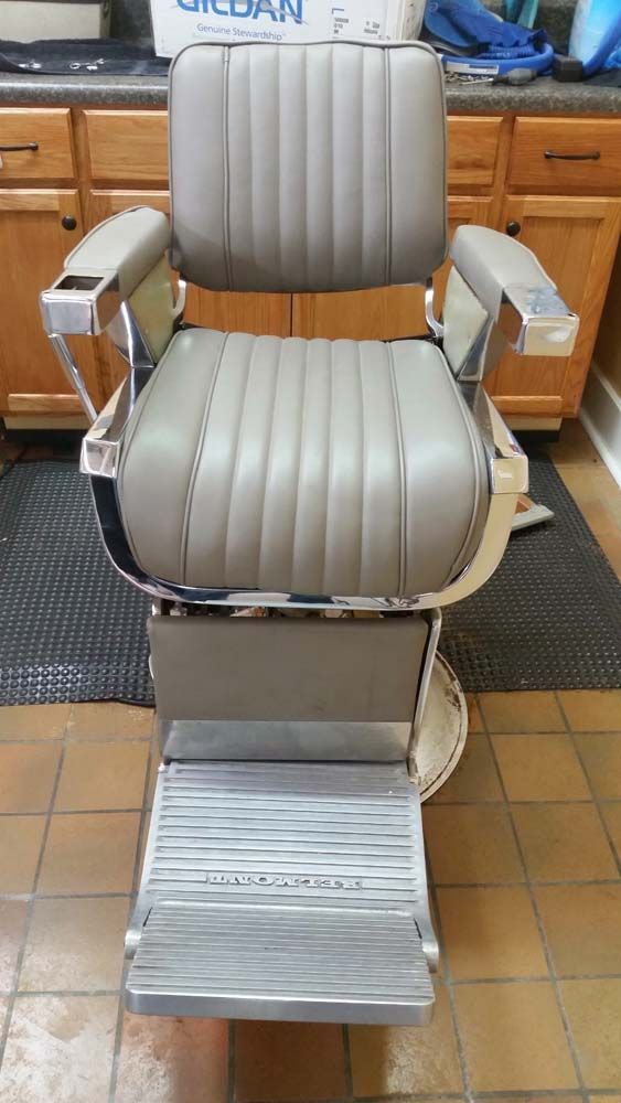 Vintage barber chair, tan upholstery, chrome frame, footrest, wooden cabinets in the background.