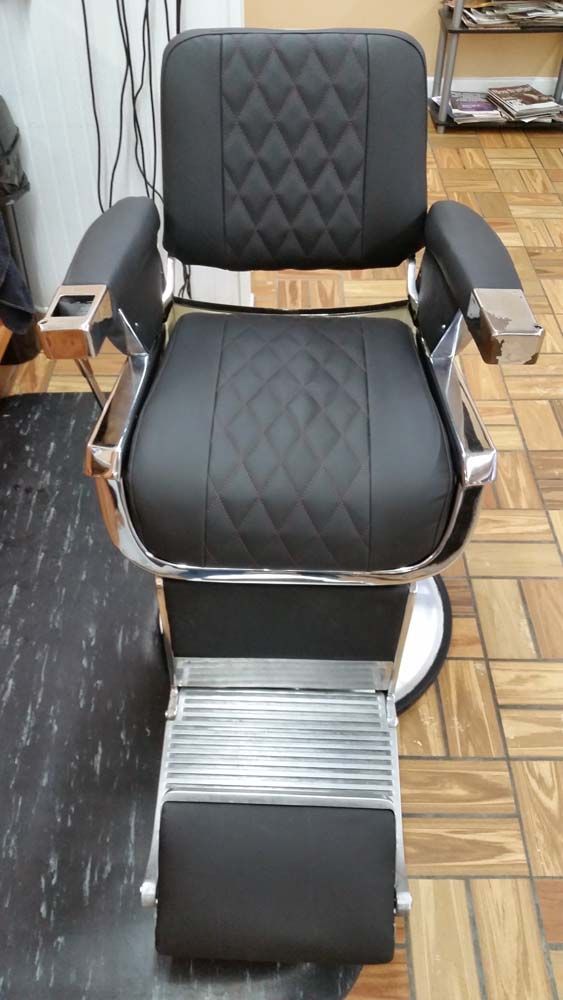 Black barber chair with diamond-stitched seat and back, chrome armrests, and footrest, in a barber shop.