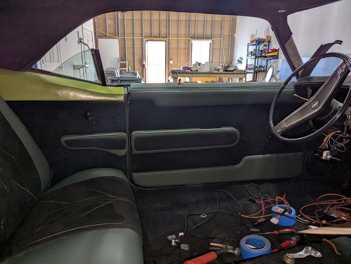 Interior of a classic car: black and green seats, door panel, steering wheel, and exposed wiring.