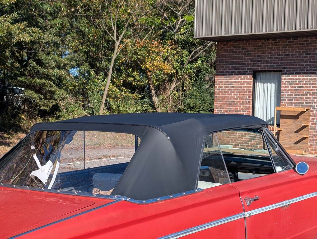 Red convertible car with a black top. It’s parked outside, with trees and a building in the background.