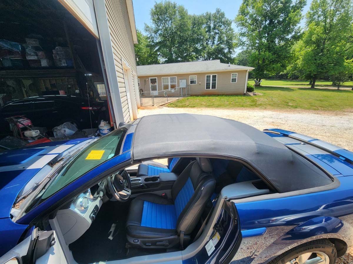 Blue convertible Mustang with black and blue seats, top down, parked outside a garage.