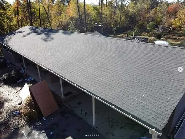 Long, gray shingle roof over a structure with white support beams, in a wooded area.
