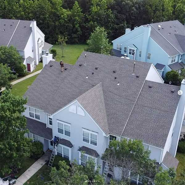 Workers on a light blue townhouse roof. Gray shingles, trees, ladders, and nearby similar homes.