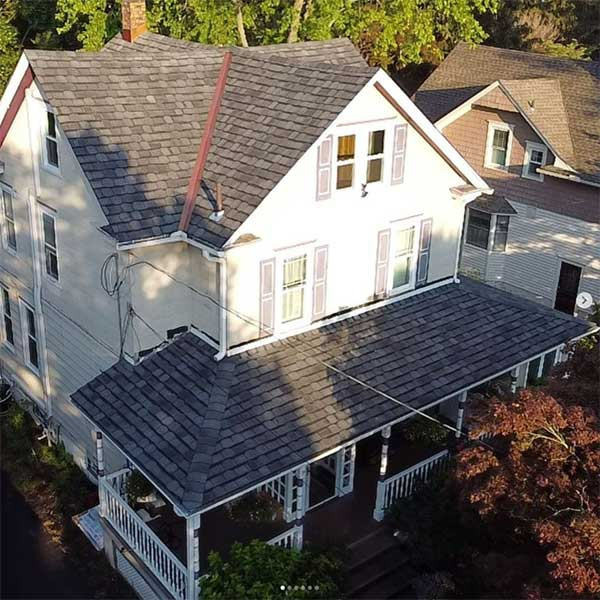 White house with gray shingle roof and porch. Another house visible in background. Trees surround.