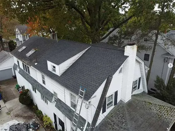 White house with dark gray roof, chimney, and dormers under a tree, with ladder on side.