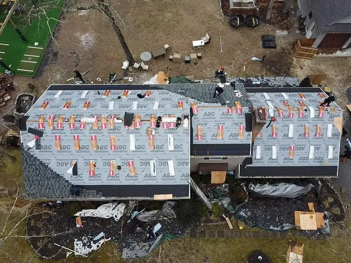 Aerial view of a house with roofing being replaced; new underlayment visible, materials and debris are scattered.