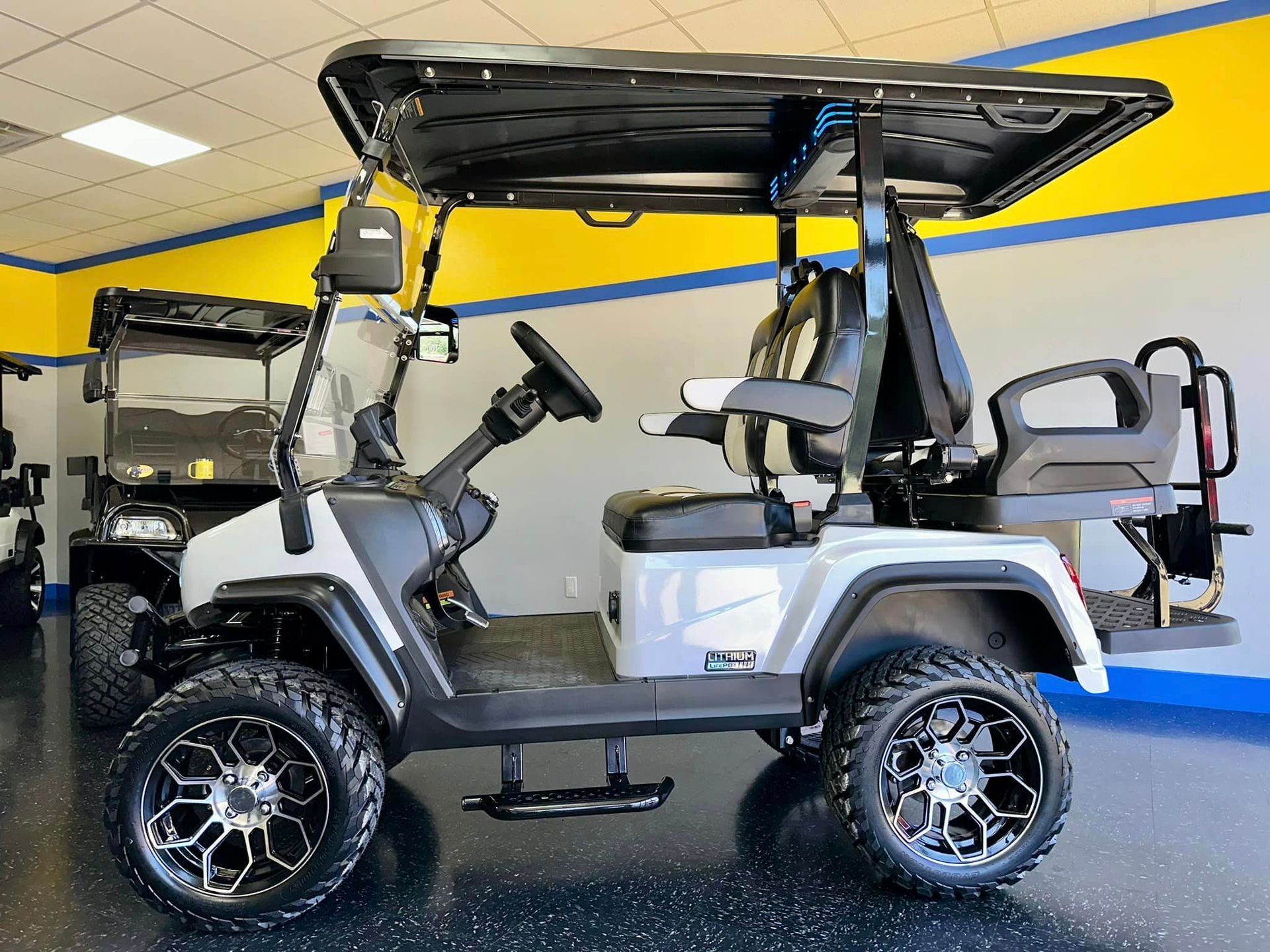 A white and black lifted golf cart with large, stylized wheels, parked in front of a yellow and blue wall.