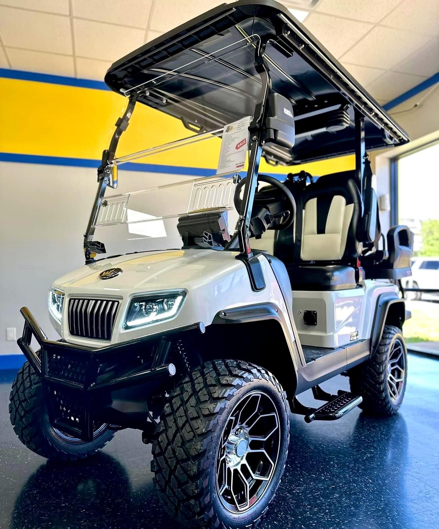 White golf cart with black accents and large, off-road tires parked indoors near a doorway. It features a custom roof and seating.
