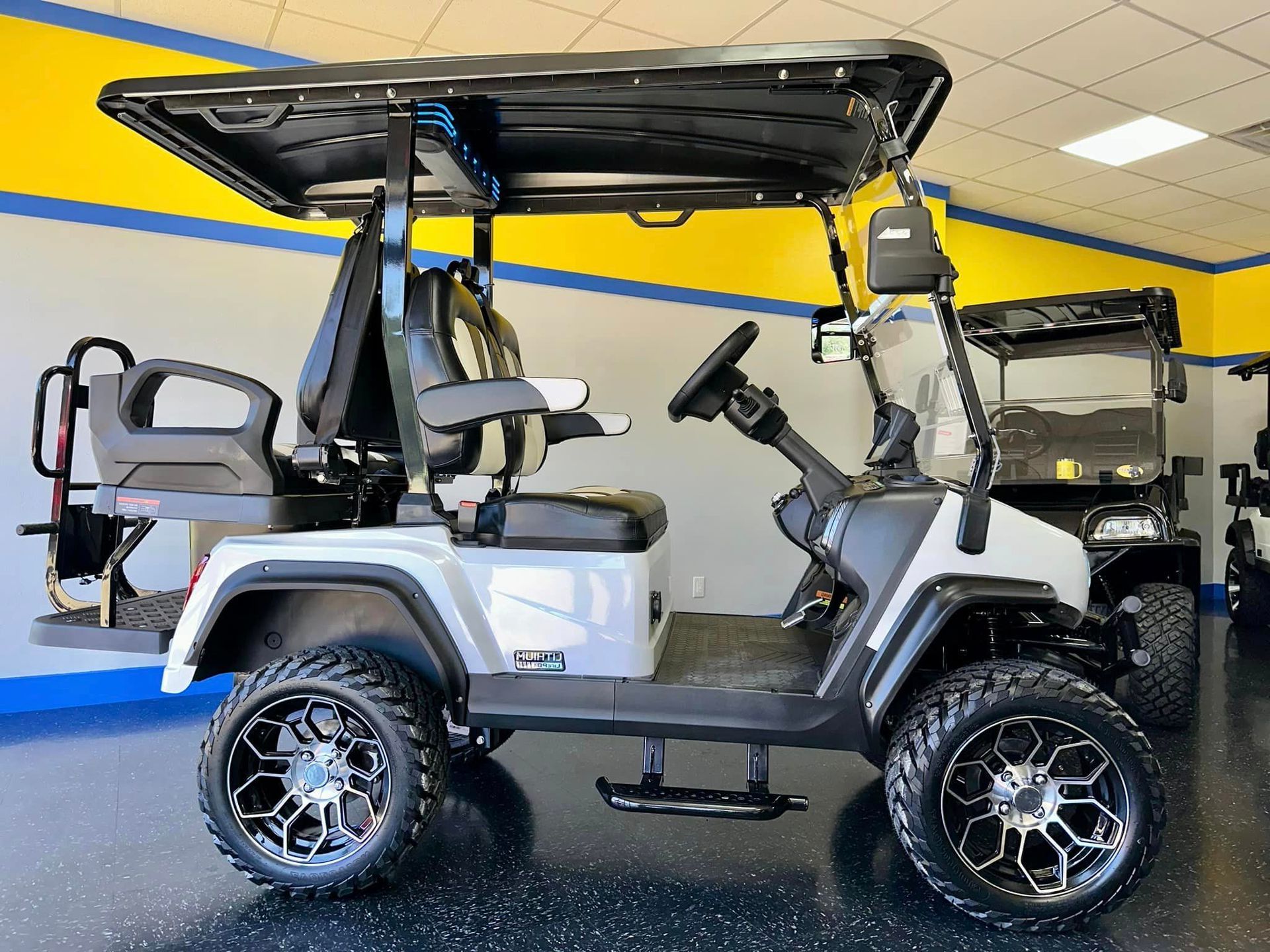 A silver golf cart with black roof, black and silver wheels, and two-tone seating, parked indoors with yellow and blue accents.