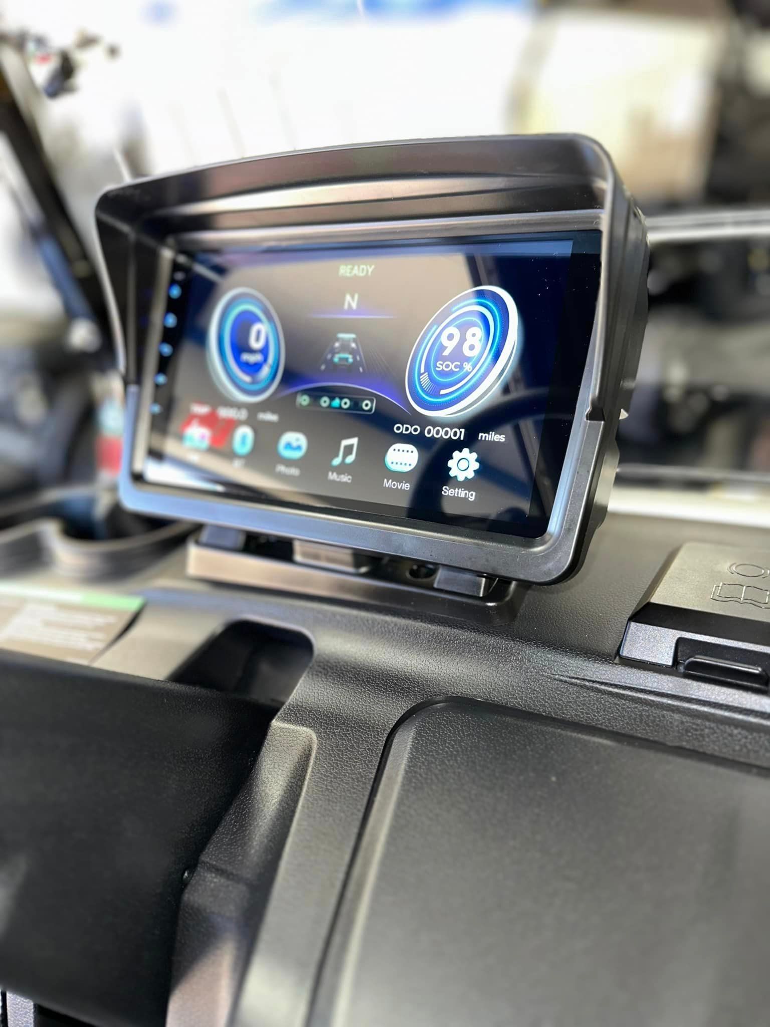 A digital dashboard display with a speedometer and gauges inside a vehicle. The screen has a black hood and is mounted on the dashboard.