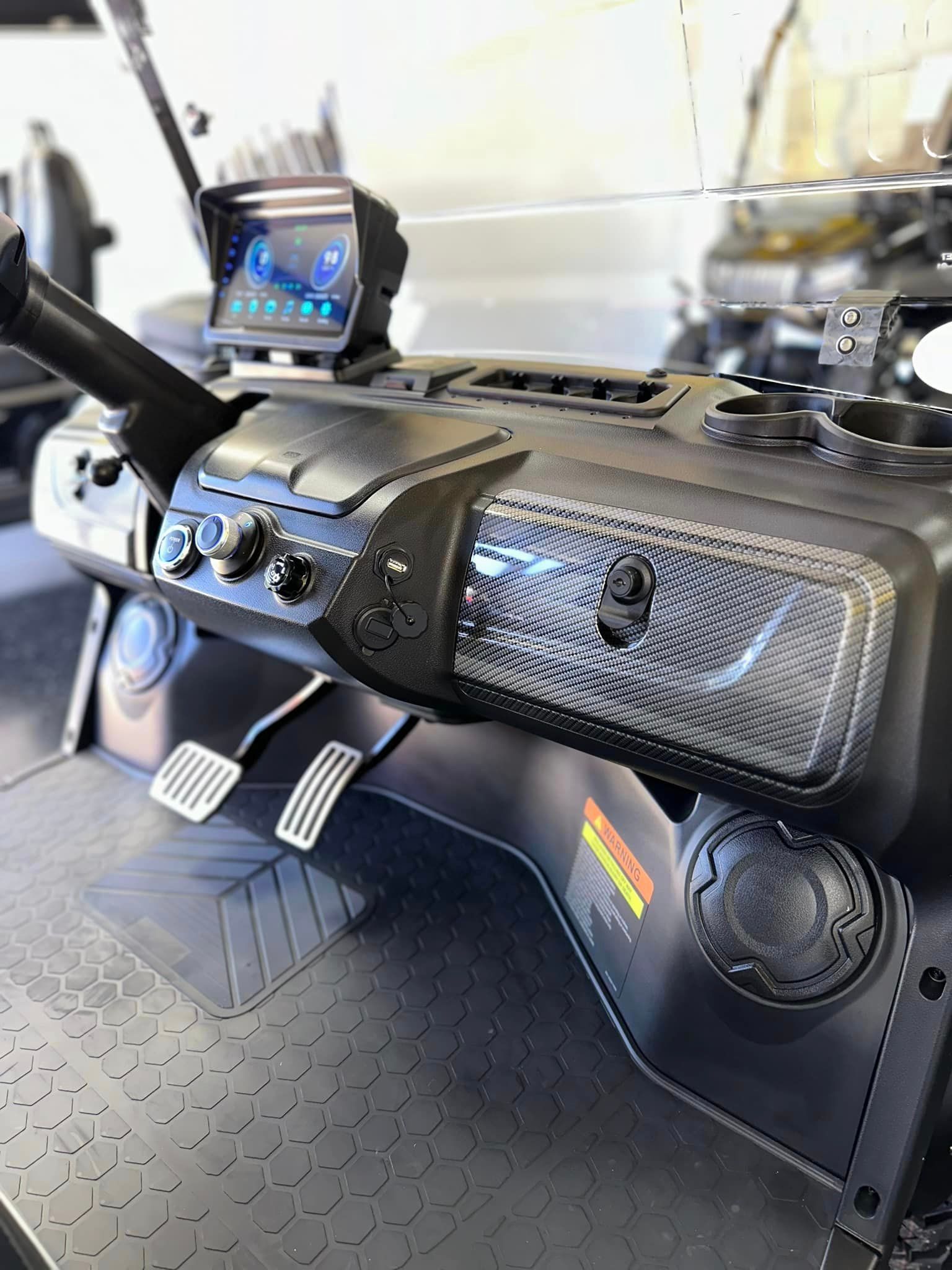 Dashboard of a utility vehicle with a digital display, controls, cup holders, and foot pedals. Black and gray colors are prominent.