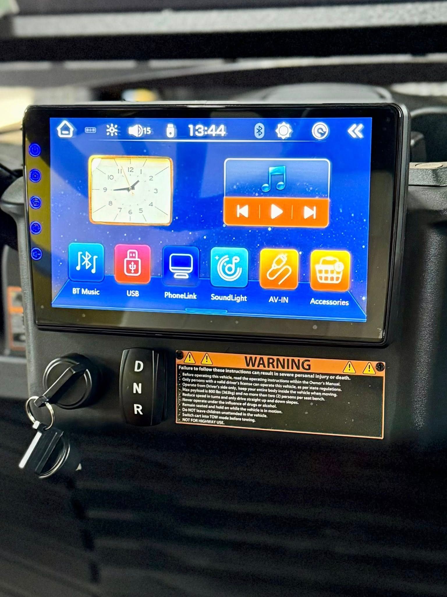 Cart dashboard with screen displaying clock, music controls, and icons; key, gear selector, and warning label.