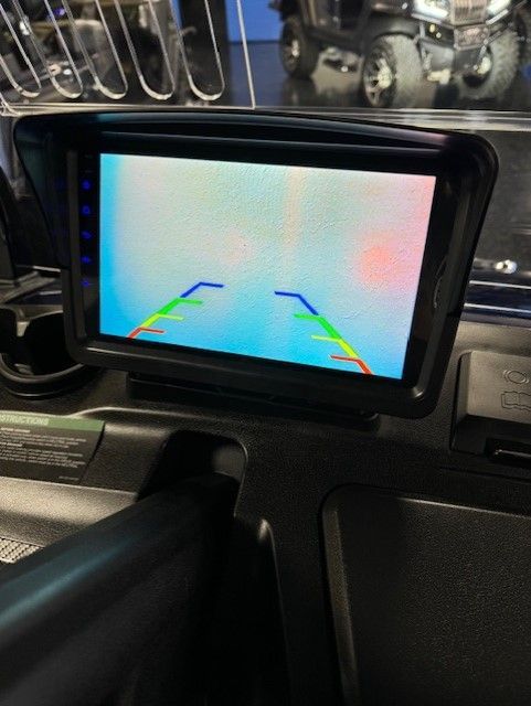 Rear-view camera monitor displaying a view behind the vehicle, with distance guidelines. Screen is mounted on dashboard.