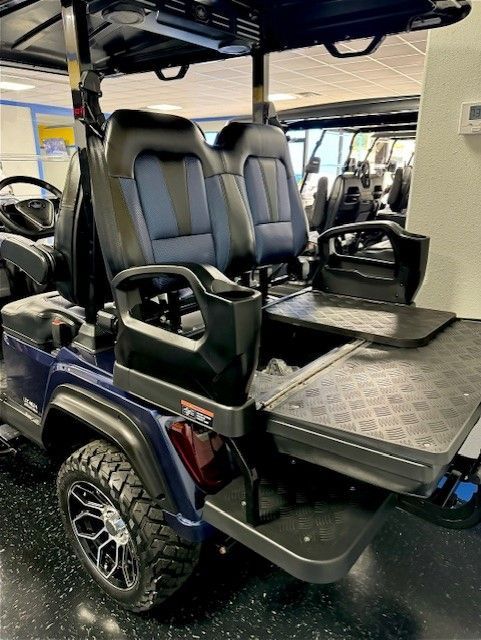 A blue golf cart with black leather seats, a rear-facing bench, and a fold-down utility surface is indoors.