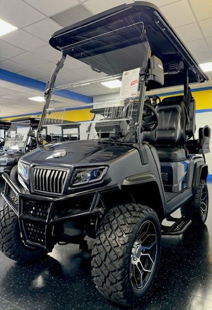 Black golf cart with rugged tires, a black grill, and black accents, displayed indoors at a dealership.