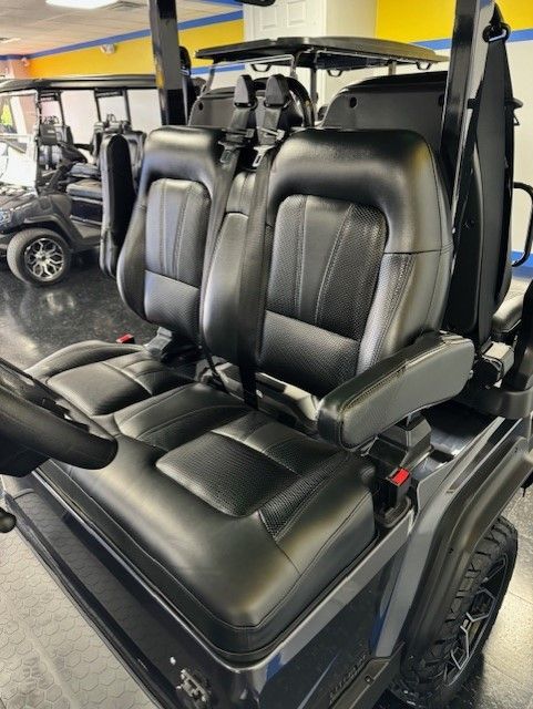 Black leather golf cart seats with armrests in a showroom. Another golf cart is visible in the background.